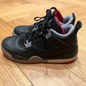 Nike Air Jordan 4 Black/Red - Säljer ett par klassiska Nike Air Jordan 4 i svart skinn med röda och grå detaljer. Skorna har meshpaneler, snörning och en ikonisk siluett med Nike-logga på hälen. Röd insida och grå sula med röd front ger en riktigt clean och sportig vibe.
