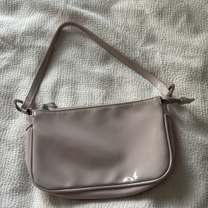 Beige axelväska i glansigt material - Snygg beige axelväska med glansig finish och silverfärgade metalldetaljer. Väskan har en enkel, stilren design med dragkedja upptill och ett rymligt svart foder inuti. Perfekt för att bära det viktigaste och ger en clean look till din outfit.