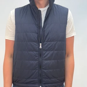 Mörkblå quiltad dunväst med dragkedja - Stilren svart dunväst med quiltad design och hög krage. Västen har hel dragkedja framtill och är lätt vadderad för extra värme. Perfekt att slänga över en hoodie eller t-shirt för en casual look.