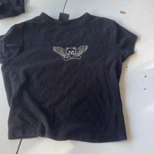 Svart topp med nalletryck från Bershka - Svart kortärmad topp från Bershka i bomull med ett tryck av en nalle med vingar på bröstet. Enkel och snygg design som passar perfekt till jeans eller kjol. Mjuk och skön att ha på sig.