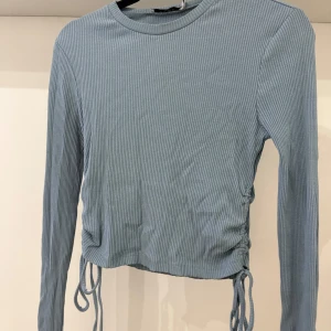 Blå långärmad topp med snörning - Säljer en ljusblå ribbad långärmad topp med snörning i sidorna. Toppen är något croppad och har en rund halsringning. Perfekt för dig som gillar en enkel men ändå trendig stil.