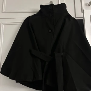 Svart poncho  - Stilren svart poncho med hög krage, knappar framtill och matchande bälte i midjan. Helt oanvänd. Ny utan prislapp.