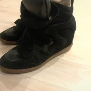 Isabel Marant svarta sneakers med kilklack - Snygga svarta sneakers från Isabel Marant med kilklack och kardborreband. Skorna har en cool, chunky design med ovandel i mocka och skinn samt en ljusbrun gummisula. Perfekta för dig som vill ha både stil och höjd i vardagen.