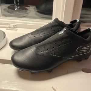 Svarta fotbollsskor RAZOR strl 39 - Säljer ett par svarta fotbollsskor från RAZOR, sketchers i storlek 39. Skorna har en clean, modern design med diskreta silverdetaljer och texten 'Carbon Infused' på sidan. Ovandelen är i syntet/textil och sulan har en cool, iriserande finish. Perfekta för dig som vill sticka ut på planen.