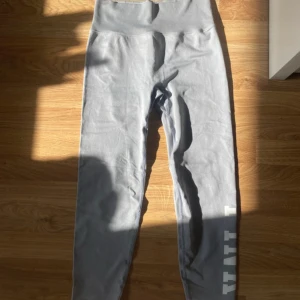 Grå leggings från PINK Active - Grå leggings från PINK Active med bred resår. Tillverkade i ett stretchigt syntetmaterial som passar perfekt för träning. Snyggt tryck med texten 'PINK' längs ena benet. Endast använd fåtal gånger så i jätte bra skick! Hör av er vid frågor!