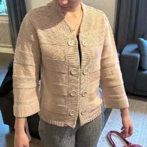 Beige stickad kofta med knappar - Snygg beige stickad kofta med bred ribbad krage och dubbla rader knappar framtill. Koftan har trekvartsärmar och ett diskret randigt mönster i stickningen. Perfekt för lager-på-lager och chillare dagar. Använd gärna köp nu