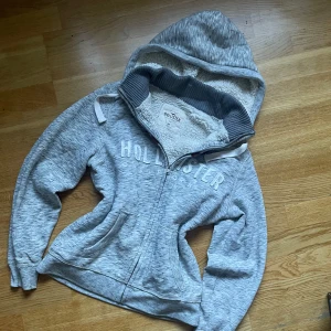 Rare y2K Hollister zip up med päls  - *   Märke: Hollister ✅ *   Storlek: M 📏 *   Färg: Grå 🎨 *   Skick: Fint skick 👍 *   Modell: Klassisk Hollister-design 🧥  Denna hoodie håller dig varm och bekväm, perfekt för alla tillfällen! 🤩 Den passar lika bra till jeans som till mjukisbyxor för en avslappnad men stilren look. Vit fluffig insida och Hollister tryckt i vitt framtill och på huvan. 👌  Missa inte chansen att lägga vantarna på denna sköna hoodie! 😉