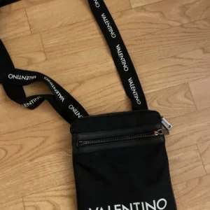 Svart axelväska från Valentino - Snygg svart axelväska från Valentino med justerbart band där loggan är tryckt i vitt. Väskan har dragkedja upptill och ett extra fack framtill med dragkedja. Tillverkad i slitstark syntet och har flera innerfack för småprylar.