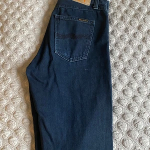 Mörkblå jeans från Nudie Jeans 28/30 - Snygga mörkblå jeans från Nudie Jeans i klassisk rak modell. Jeansen har fem fickor, subtila sömdetaljer på bakfickan och är tillverkade i mjukt denimtyg av bomull. Perfekta för dig som gillar stilrena och tidlösa jeans.