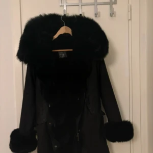 Svart pälsjacka med stor krage - Säljer en svart pälsjacka med stor fluffig krage och breda pälskanter vid ärmsluten. Jackan har en klassisk och lyxig vibe, perfekt för kalla dagar när du vill sticka ut. Stängs med knappar framtill och har en snygg siluett.