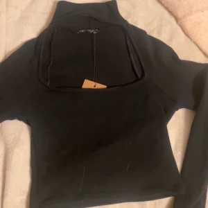 Svart långärmad polotopp - Säljer en svart långärmad polotopp med hög krage och en cool öppen detalj framtill. Toppen är croppad och har en stilren, minimalistisk look som passar perfekt till jeans eller kjol. Materialet känns mjukt och stretchigt.