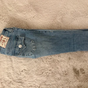 True Religion Joey Low Rise Flare Jeans - Snygga ljusblå jeans från True Religion, modell Joey Low Rise Flare. Klassisk femficksdesign med markerade sömmar och ikonisk bakficka med lock och knapp. Jeansen har låg midja och utsvängda ben för en cool retrostil. Perfekta för dig som gillar statement-jeans.