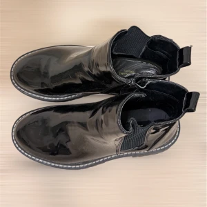 Svarta Ankel  boots - Säljer ett par svarta Ankel boots i glansigt lackmaterial med bred resår på sidorna och kraftig platt sula. Skorna har rund tå och dragflikar bak för enkel påtagning. Perfekta för dig som vill ha en edgy och stilren look.
