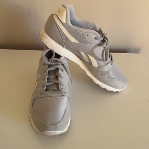 Gråa sneakers med vita detaljer från Reebok - Snygga grå sneakers från Reebok med vita detaljer och logga på sidan och hälen. Skorna har snörning, rund tå och platt sula. Ovandelen är i en mix av syntet och mocka för en klassisk look. Använda två tillfällen, se bilderna. Storlek 40. I fint skick förutom att de är lite smutsiga. Annars är de hela och fina, inga slitage och anmärkningar. 