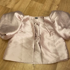 Ljusrosa satintopp med puffärm Gina Tricot - Superfin ljusrosa blus från Gina Tricot i satinliknande material. Toppen har stora puffärmar och knytning framtill med två rosetter. Perfekt för dig som vill sticka ut med en söt och trendig look.