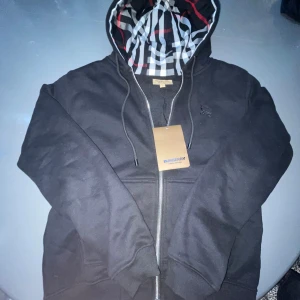 Svart Burberry hoodie med rutig huva - Svart hoodie från Burberry med dragkedja och klassisk rutig huva i svart, vitt och rött. Broderad logga på bröstet och snörning i huvan. Perfekt för dig som vill ha en stilren och lyxig streetwear-look.