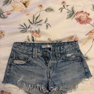 Blå jeansshorts från Pull&Bear - Snygga ljusblå jeansshorts från Pull&Bear med låg midja, slitna detaljer och fransig kant. Klassisk femficksmodell med knappar framtill och bakfickor med knapp. Perfekta för sommaren och ger en cool, avslappnad vibe.