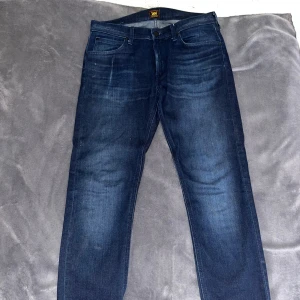 Mörkblå jeans från Lee - Säljer ett par mörkblå jeans från Lee med klassisk femficksdesign och snygg tvätt. Jeansen har Lee-logga på bakfickan och är tillverkade i mjukt denimtyg. Passar perfekt till en avslappnad stil och har en rak passform. Modell: LUKE. Storlek: 31/32