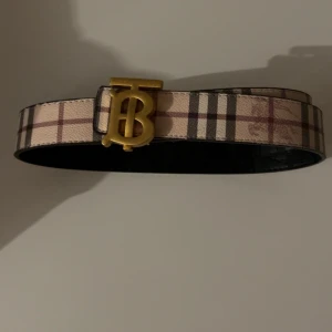 Burberry skärp med guldspänne - Snyggt bälte med klassiskt rutigt mönster i beige, brunt och svart. Bältet har ett stort guldigt spänne med Burberrys ikoniska TB-logga. Perfekt accessoar för att lyfta din outfit med en lyxig touch.