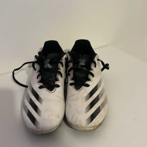 Vita Adidas fotbollsskor med svarta detaljer - Säljer ett par vita Adidas fotbollsskor med svarta ränder och grafiskt mönster på sidorna. Skorna har snörning och platt sula med dobbar för bra grepp på planen. Perfekta för dig som vill sticka ut på matchen!