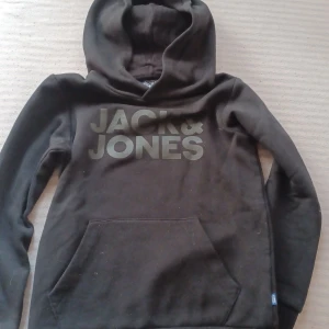 Svart barn hoodie från Jack & Jones - Svart barn  hoodie från Jack & Jones med stor logga framtill. Klassisk känguruficka och huva. Mjuk insida och långärmad modell, perfekt för kyliga dagar. Snygg och enkel design som funkar till det mesta...Storlek 140.