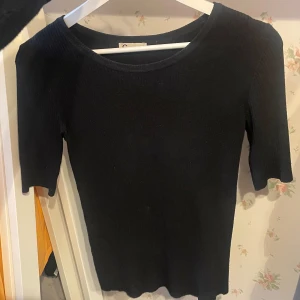 Svart ribbad topp från Cubus - En svart ribbad topp från Cubus i storlek XS. Toppen har rund halsringning och halvlånga ärmar. Den är figurnära och tillverkad i ett stretchigt material som sitter snyggt på kroppen. Perfekt att styla med jeans eller kjol för en clean look.