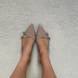 Beiga lackade pumps med spetsig tå - Snygga pumps i beige lack med spetsig tå och låg klack. Skorna har två smala remmar med silverspännen över foten och öppen häl med justerbart spänne. Perfekta för dig som gillar en chic och stilren look.