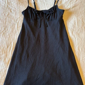 Svart klänning från Gina Tricot - Snygg svart klänning från Gina Tricot med smala axelband och knytning framtill. Perfekt för en stilren look. Klänningen har en enkel och elegant design som passar till många tillfällen.