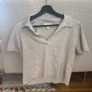 Beige ribbad topp med krage från H&M - Säljer en grå ribbad topp / tshirt från H&M i storlek M. Toppen har korta ärmar, klassisk krage och v-ringning. Materialet är mjukt och stretchigt, perfekt för en chill och stilren look. Passformen är normal och toppen är enkel att matcha med jeans eller kjol.