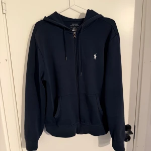 Marinblå hoodie från Polo Ralph Lauren - Snygg marinblå hoodie från Polo Ralph Lauren med dragkedja framtill, huva med snörning och klassisk vit logga på bröstet. Mjuk bomullskvalitet och två fickor framtill. Perfekt för chill dagar eller när du vill ha en clean look.