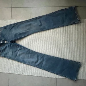 Blå bootcut jeans med rå kant - Säljer ett par blå jeans med bootcut-modell och rå, ofållad kant längst ner. Jeansen har klassisk femficksdesign och knappgylf. Perfekta för dig som gillar en avslappnad och trendig look.
