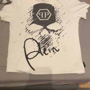 Vit t-shirt från Philipp Plein med tryck - Snygg vit t-shirt från Philipp Plein med stort svart döskalletryck och logga på bröstet. T-shirten har korta ärmar, rund hals och är gjord i mjuk bomull. Märkesetikett och logga även bak i nacken. Perfekt för dig som gillar streetstyle.