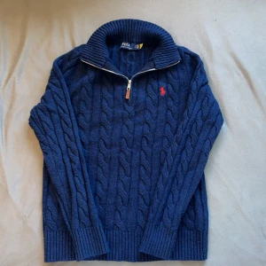 Quarter Zip Polo Ralph Lauren - Kabelstickad marinblå tröja från Polo Ralph Lauren med halv dragkedja och hög krage. Tröjan har lång ärm, ribbade muddar och röd broderad logga på bröstet. Perfekt för lager-på-lager och snygg till jeans eller chinos. Perfekt inför vintern.