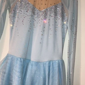 Ljusblå glittrig klänning med strass - Superfin ljusblå konståkningsklänning med långärmade meshärmar och massor av glittrande strassdetaljer över hela. Bara att trä på och se ut som en riktig isprinsessa 😍 Älskar denna så himla mycket men kommer tyvär inte för användning längre.
