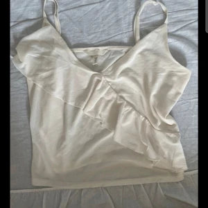 Vit linnetopp med volang från H&M - Snygg vit linnetopp från H&M med smala axelband och v-ringning. Toppen har en dekorativ volang som går diagonalt över framsidan och är tillverkad i ett mjukt, lätt material. Perfekt för en fräsch och stilren look.