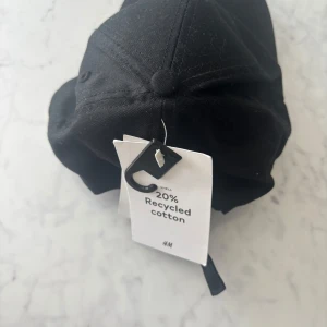 Svart keps från H&M i bomull - Svart klassisk keps från H&M med böjd skärm och justerbar rem baktill. Tillverkad i bomullstyg med 20% återvunnen bomull, perfekt för en clean och stilren look. Passar dig som gillar enkel och tidlös streetstyle.