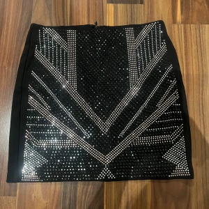 Svart glittrig kortkjol med paljetter - Snygg svart kortkjol med glittriga paljettdetaljer i geometriskt mönster framtill. Kjolen har en tight passform och dragkedja bak. Perfekt för dig som vill sticka ut med en cool och festlig look.