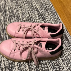 Adidas Stan Smith rosa sneakers - Säljer ett par rosa Adidas Stan Smith sneakers med svart detalj på hälen och gummisula. Skorna har klassisk snörning och är tillverkade i skinn. Perfekta för dig som vill sticka ut med färg på fötterna!