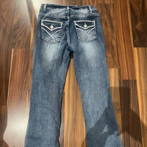 Blå bootcut jeans med detaljerade fickor - Snygga blå jeans med bootcut passform och coola detaljer på bakfickorna. Jeansen har ljusa slitningar framtill och bak, klassisk femficksdesign och kontrastsömmar. Perfekta för dig som gillar en avslappnad men trendig look.