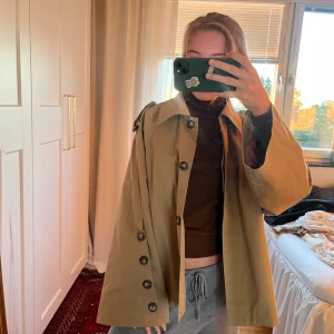 Beige kort trenchcoat med knappar - Säljer en beige kort trenchcoat med stora knappar framtill och på ärmarna. Jackan har klassisk krage, axelklaffar och en loose passform. Perfekt för lager-på-lager och enkel att styla till olika outfits.