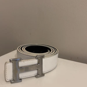 Vitt skärp med H-spänne i skinn - Snyggt vitt bälte i skinn med ett stort, silvrigt H-format spänne. Bältet har svart insida och stilrena sömmar längs kanterna. Perfekt accessoar för att lyfta din outfit med en clean och modern look. Har gjort två egna hål för bälte va för stort för mig