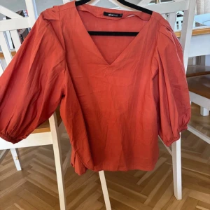 Rostfärgad blus från Gina Tricot - Säljer en snygg rostfärgad blus från Gina Tricot med v-ringning och puffiga trekvartsärmar med resår vid ärmslut. Blusen har en lös passform och är tillverkad i ett lätt bomullstyg, perfekt för vår och sommar.