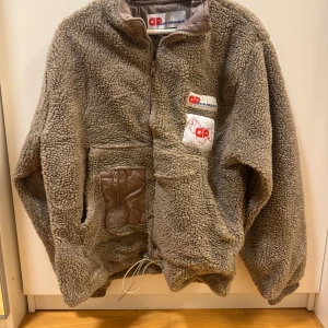 Beige teddyjacka från Denim Project - Mysig beige teddyjacka från Denim Project med dragkedja framtill, två bröstfickor med logga och en ficka med dragkedja. Jackan har hög krage, snörning nedtill och detaljer i syntetmaterial. Perfekt för kalla dagar.