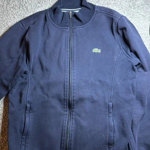 Marinblå sportjacka från Lacoste - Snygg marinblå sportjacka från Lacoste med dragkedja framtill och klassisk krokodil-logga på bröstet. Jackan har lång ärm, ståkrage och är tillverkad i mjuk bomull med fleecefoder på insidan. Perfekt för en sportig och clean look. Den är använd men inget dåligt skick. Är dock rätt så liten passar om du är 160-172