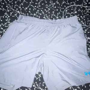 Grå träningsshorts från Under Armour - Snygga grå träningsshorts från Under Armour med elastisk midja och blå logga på benet. Modellen är loose fit och tillverkad i ett lätt och snabbtorkande syntetmaterial, perfekt för intensiva träningspass eller chill dagar.