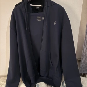 Marinblå hoodie från Polo Ralph Lauren - Säljer en marinblå hoodie från Polo Ralph Lauren med klassisk broderad logga på bröstet. Tröjan har huva med snörning, dragkedja framtill och två fickor. Perfekt för dig som gillar stilren och sportig look. Har inget kvitto på den.