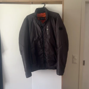 Svart Sail Racing pufferjacka herr - Säljer en svart pufferjacka från Sail Racing med orange foder och snygga logodetaljer på bröstet och ärmen. Jackan har dragkedja framtill och är tillverkad i lätt syntetmaterial med Primaloft-foder. Perfekt för höst och vinter, stilren och sportig.  JACKAN ÄR VÄLDIGT SVÅR ATT FÅ TAG I 