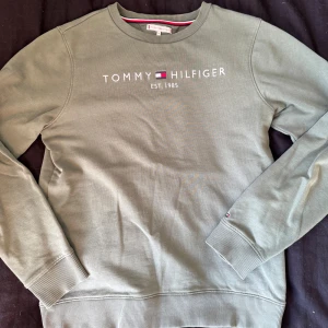 Grön Tommy Hilfiger sweatshirt - Snygg grön sweatshirt från Tommy Hilfiger med broderad logga och text på bröstet. Tröjan har rund hals, långa ärmar och ribbade muddar vid ärmslut och nederkant. Perfekt för en chill och stilren look.