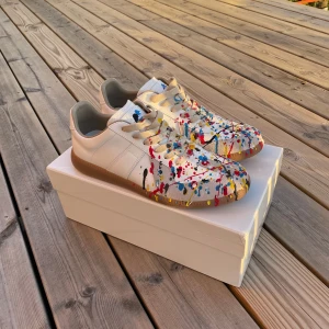 Maison Margiela sneakers paint splatter - Maison Margiela sneakers i vitt skinn med beige mockadetaljer. Unik paint splatter-design i rött, blått, gult och svart över hela skon. Perfekt statement piece. Hör gärna av er för fler bilder eller funderingar. Köpta begagnade