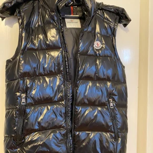 Svart dunväst från Moncler - Snygg svart dunväst från Moncler med glansig finish och klassisk logga på bröstet. Västen har huva, två dragkedjefickor och quiltad design. Perfekt för lager-på-lager och streetstyle. Stängs med dragkedja framtill.